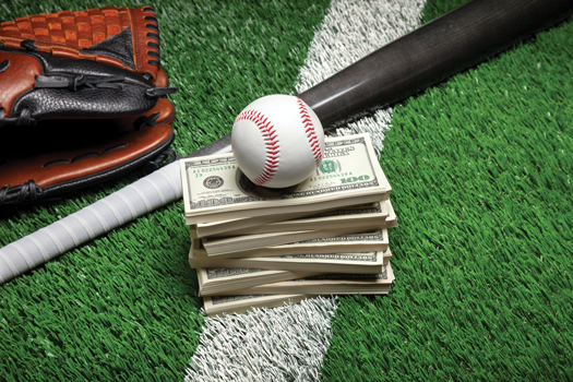 A stack of hundred-dollar bills on a baseball field with a baseball resting on top. Nearby, a leather baseball glove and a wooden bat lie on the artificial turf, symbolizing the financial aspects of professional sports.