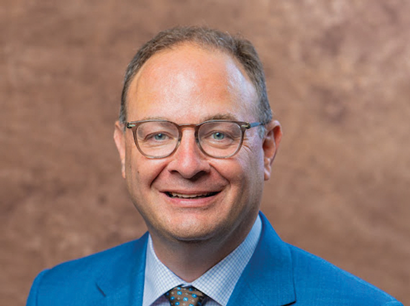 A man with short, light brown hair, glasses, and a friendly smile. He is wearing a blue suit, white dress shirt, and a patterned tie. The background is a textured brown surface.